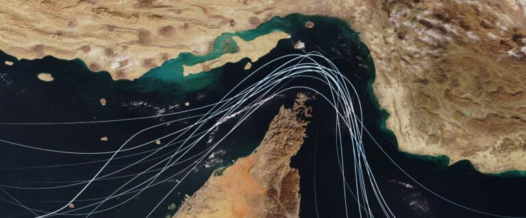 Satellite view of the Strait of Hormuz with white graphic lines representing global shipping lanes and maritime traffic between the Persian Gulf and Gulf of Oman. Strategic oil transport concept